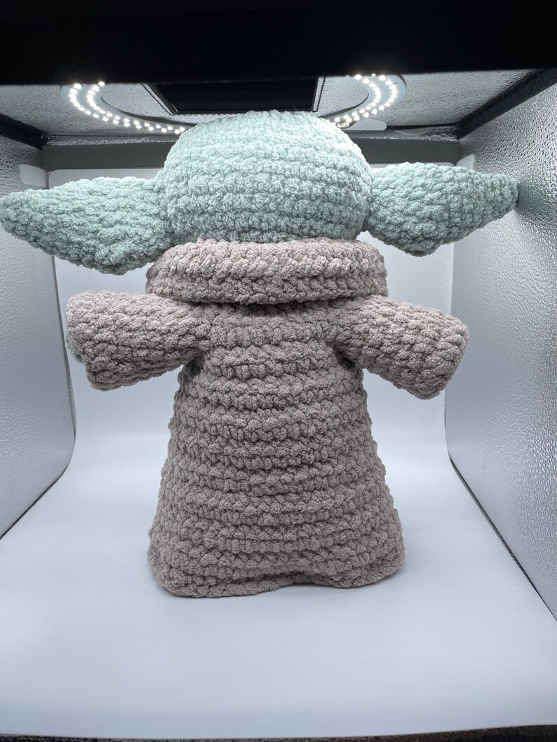 May include: A crocheted plush toy resembling a character from the Star Wars franchise. The toy is wearing a brown hooded robe and has a green head and ears.