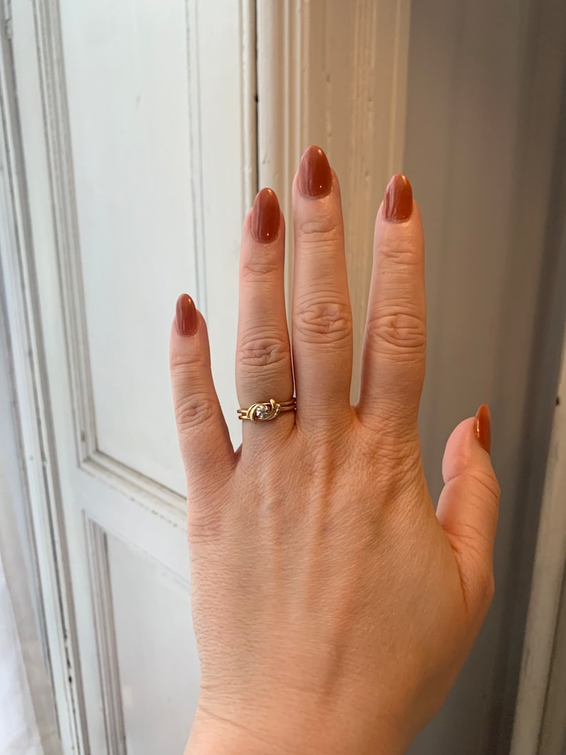May include: A gold ring with a small diamond set in a twisted band. The ring is worn on the ring finger of a hand with brown nail polish.