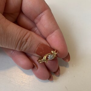 May include: A gold ring with a single diamond in the center. The ring is designed with two intertwined bands that form a swirl pattern.