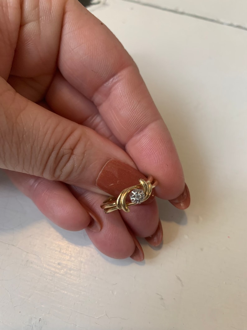 May include: A gold ring with a single diamond in the center. The ring is designed with two intertwined bands that form a swirl pattern.