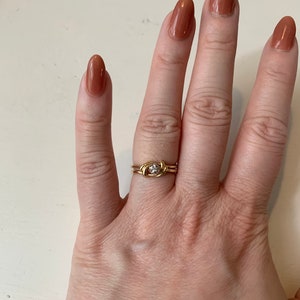 May include: A gold ring with a small, clear stone set in the center. The ring is on a finger with brown nail polish.