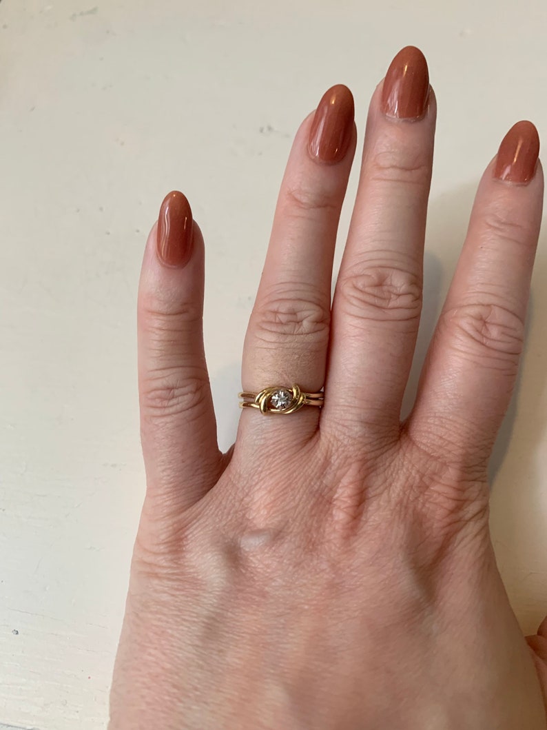 May include: A gold ring with a small, clear stone set in the center. The ring is on a finger with brown nail polish.