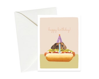 May include: A birthday card featuring a dachshund wearing a purple party hat and holding a lit candle, peeking out of a hot dog bun. The card has the text "happy birthday!" in orange.
