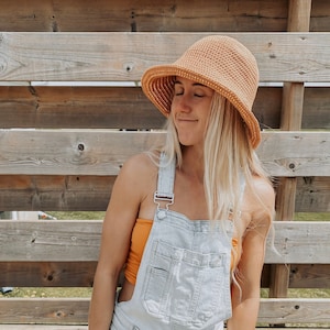 Op de afbeelding: Een vrouw draagt een lichtblauwe denim overalljurk met een lichtoranje gehaakte hoed. De hoed heeft een brede rand en is gemaakt van een zacht, getextureerd materiaal. De vrouw staat voor een houten hek.