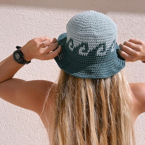 Può includere: Una persona indossa un cappello a secchiello in crochet verde e blu con un design bianco. Il cappello ha un'ampia tesa ed è realizzato con un filato morbido e testurizzato.