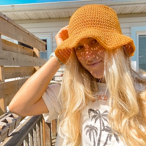 Op de afbeelding: Een vrouw met lang blond haar draagt een gele gehaakte hoed in de vorm van een emmer. Ze draagt een wit T-shirt met een palmboomprint.