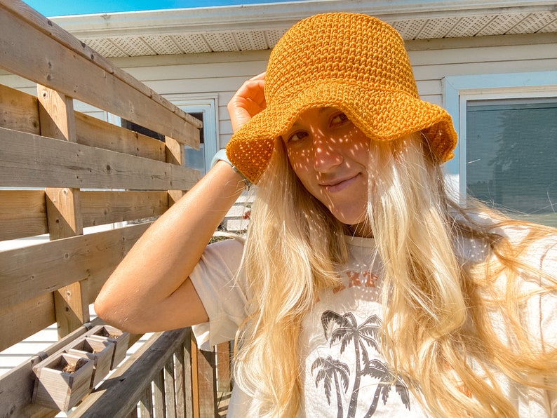 Op de afbeelding: Een vrouw met lang blond haar draagt een gele gehaakte hoed in de vorm van een emmer. Ze draagt een wit T-shirt met een palmboomprint.