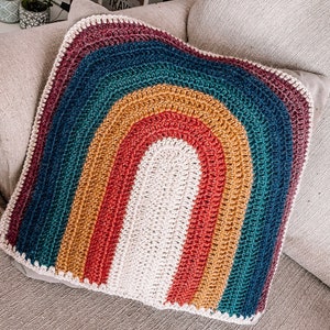 May include: A crocheted rainbow pillow with a textured, multi-colored design. The pillow is made of yarn in shades of blue, green, yellow, orange, and red.