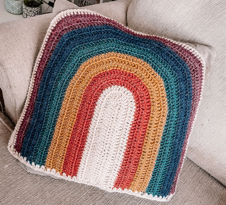 May include: A crocheted rainbow pillow with a textured, multi-colored design. The pillow is made of yarn in shades of blue, green, yellow, orange, and red.