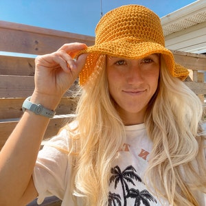 Op de afbeelding: Een vrouw draagt een gele gehaakte bucket hat met een brede rand. Ze draagt een wit t-shirt met een palmboomprint en de tekst "Just Me".