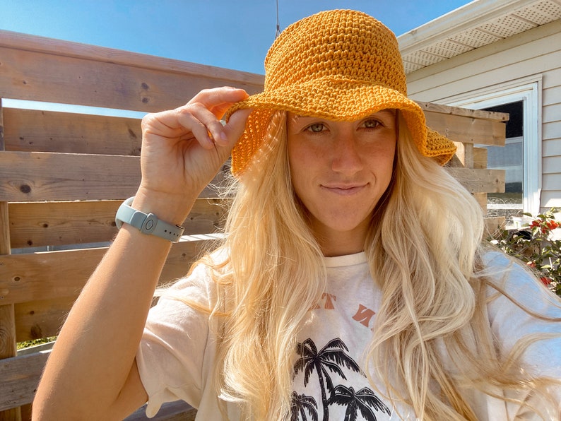 Op de afbeelding: Een vrouw draagt een gele gehaakte bucket hat met een brede rand. Ze draagt een wit t-shirt met een palmboomprint en de tekst "Just Me".