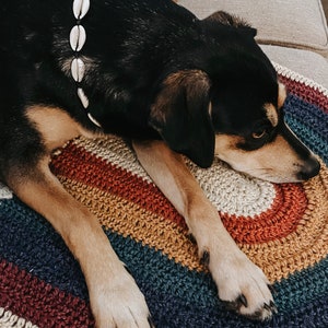 May include: A black and brown dog with a white shell necklace is lying on a multi-colored crocheted rainbow rug.