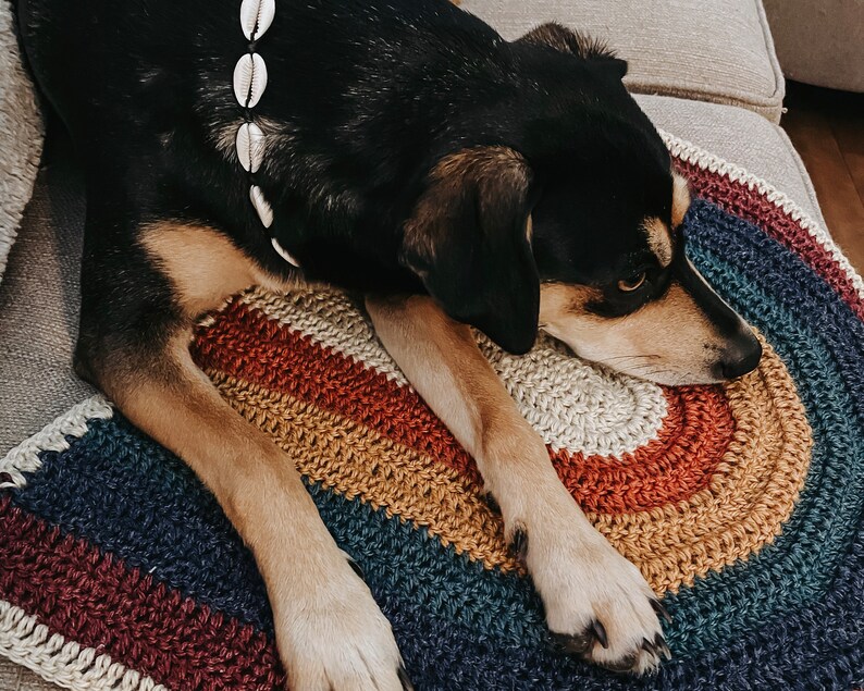 May include: A black and brown dog with a white shell necklace is lying on a multi-colored crocheted rainbow rug.