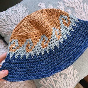 May include: A handmade crochet bucket hat in shades of tan, light blue, and navy blue. The hat features a geometric pattern in light blue and tan on the crown, transitioning to a solid navy blue brim. The hat is shown on a light blue pillow.