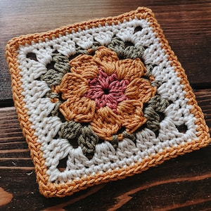 K&ouml;nnte beinhalten: Quadratischer geh&auml;kelter Untersetzer mit einem zentralen Blumendesign in Orange und Rosa, umgeben von gr&uuml;nen Bl&auml;ttern. Der Untersetzer hat einen wei&szlig;en Hintergrund und einen orangefarbenen Rand, ideal f&uuml;r die Heimdekoration.
