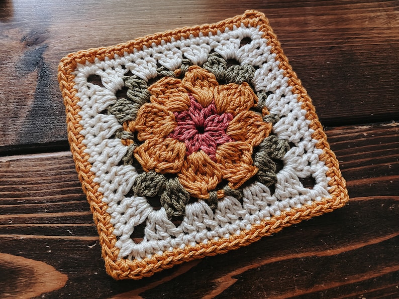 K&ouml;nnte beinhalten: Quadratischer geh&auml;kelter Untersetzer mit einem zentralen Blumendesign in Orange und Rosa, umgeben von gr&uuml;nen Bl&auml;ttern. Der Untersetzer hat einen wei&szlig;en Hintergrund und einen orangefarbenen Rand, ideal f&uuml;r die Heimdekoration.