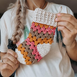 May include: A crocheted stocking with a white, brown, pink and orange colour pattern. The stocking is made with a granny square stitch.