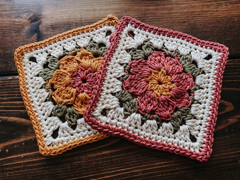 K&ouml;nnte beinhalten: Zwei quadratische H&auml;keluntersetzer. Ein Untersetzer hat eine mittige Blume in Orange- und Rosat&ouml;nen, umgeben von gr&uuml;ner und wei&szlig;er H&auml;kelarbeit. Der andere Untersetzer hat eine rosa Mittelblume mit gr&uuml;ner und wei&szlig;er H&auml;kelarbeit. Beide haben eine quadratische Form.