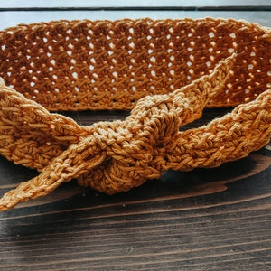 May include: Mustard yellow crochet headband with a knotted bow detail. The headband is made of a textured yarn and has an open weave design. The headband is shown on a dark wood surface.
