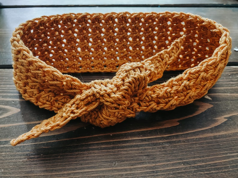 May include: Mustard yellow crochet headband with a knotted bow detail. The headband is made of a textured yarn and has an open weave design. The headband is shown on a dark wood surface.