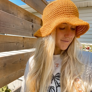 Op de afbeelding: Een vrouw met lang blond haar draagt een gehaakte bucket hat in mosterdgeel. De hoed heeft een brede rand en een textuurontwerp.