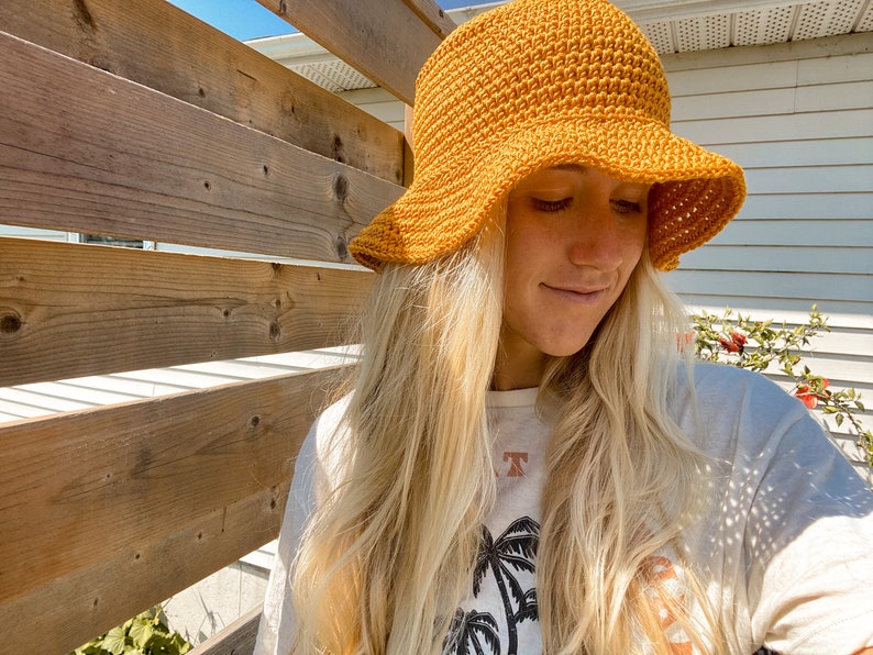Op de afbeelding: Een vrouw met lang blond haar draagt een gehaakte bucket hat in mosterdgeel. De hoed heeft een brede rand en een textuurontwerp.