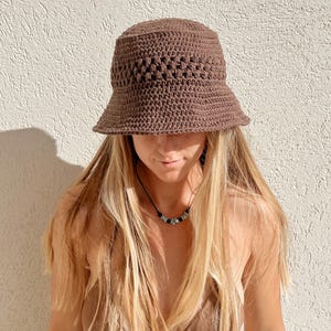 May include: A brown crochet bucket hat with a wide brim. The hat is made of a textured yarn and has a slightly open weave pattern on the crown. The hat is a solid color and appears to be handmade.