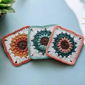 May include: Three crocheted square coasters with a floral design in shades of orange, green, and pink. The coasters are made with white yarn and have a border of the same colour as the centre flower.