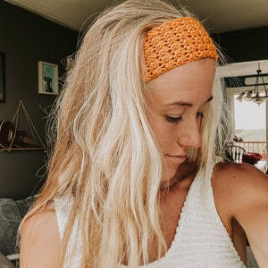 May include: An orange crochet headband. The wide headband is made of a textured, woven material. The woman in the image is wearing a white top and has long blonde hair. The background includes framed pictures and a ukulele.