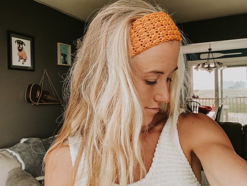 May include: An orange crochet headband. The wide headband is made of a textured, woven material. The woman in the image is wearing a white top and has long blonde hair. The background includes framed pictures and a ukulele.