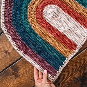 May include: A rainbow crochet rug with a white center oval. The rug is made with different colored yarn, including purple, teal, mustard yellow, and red.