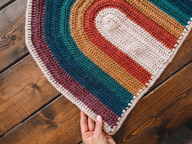 May include: A rainbow crochet rug with a white center oval. The rug is made with different colored yarn, including purple, teal, mustard yellow, and red.