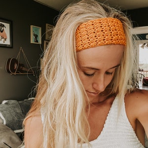 May include: A bright orange crocheted headband is worn across the forehead. The headband has a textured, open-weave design. The person in the image has long, blonde hair and is wearing a white top. The background includes framed artwork and a guitar.