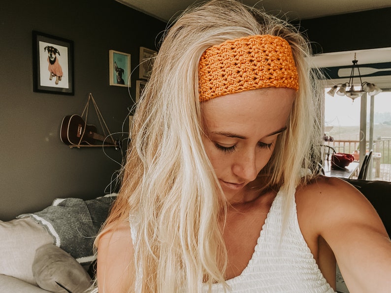 May include: A bright orange crocheted headband is worn across the forehead. The headband has a textured, open-weave design. The person in the image has long, blonde hair and is wearing a white top. The background includes framed artwork and a guitar.