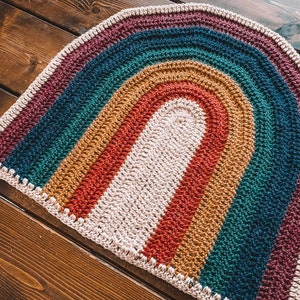 May include: A crocheted rainbow rug with a cream-colored center and a burgundy, teal, mustard, and red striped pattern.