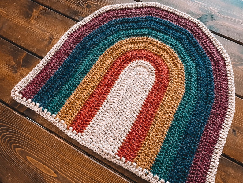 May include: A crocheted rainbow rug with a cream-colored center and a burgundy, teal, mustard, and red striped pattern.