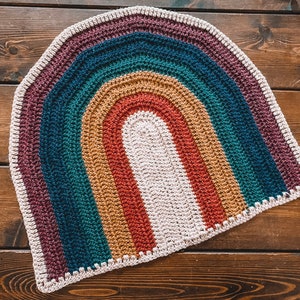 May include: A crocheted rainbow rug with a white center and a brown wooden background.