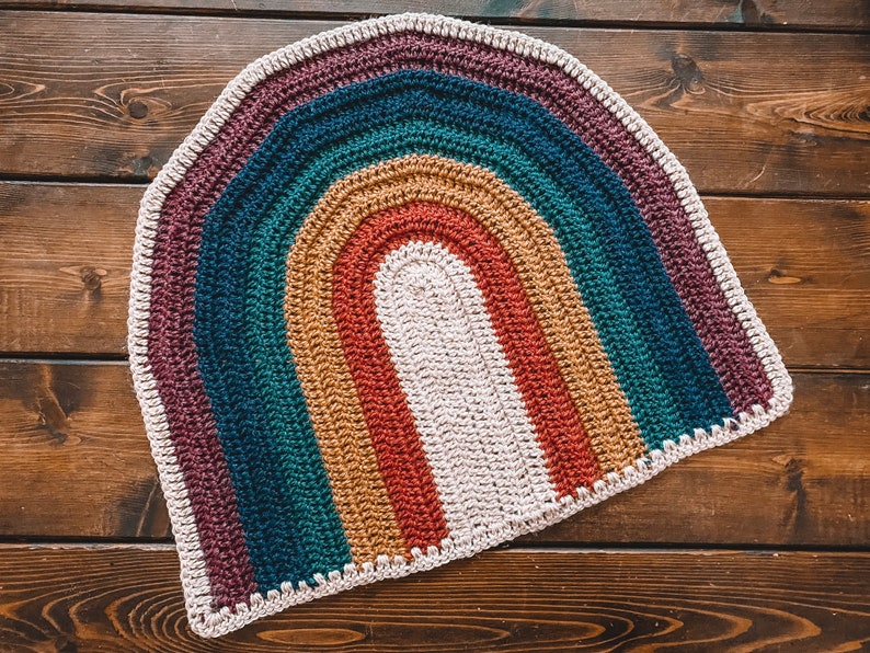 May include: A crocheted rainbow rug with a white center and a brown wooden background.
