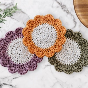 May include: Three crocheted coasters in different colors: purple, orange, and green. Each coaster has a white center and a scalloped edge.