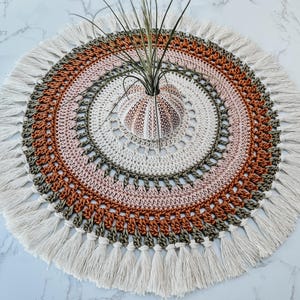 May include: A round crocheted table runner with a fringe border in shades of brown, green, pink, and white. The runner has a central circle with a white crocheted design.