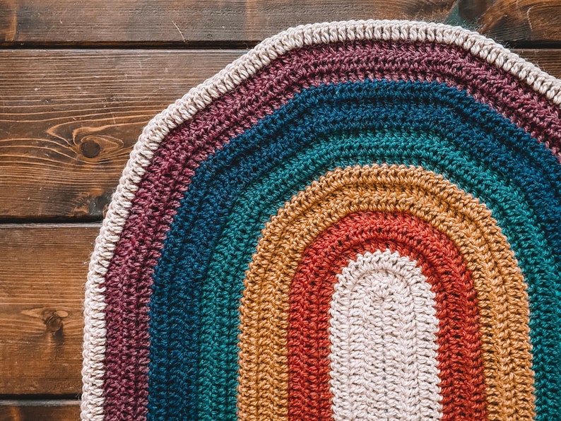 May include: A colorful crocheted oval rug with a rainbow pattern in shades of purple, teal, mustard yellow, red, and white.