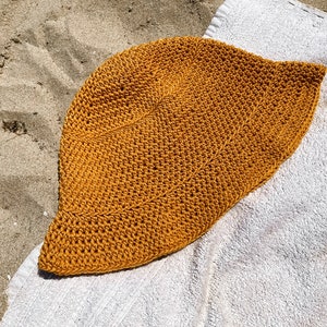 Op de afbeelding: Een gehaakte zonnehoed in een mosterdgele kleur. De hoed is gemaakt van dik, getextureerd garen en heeft een brede rand.