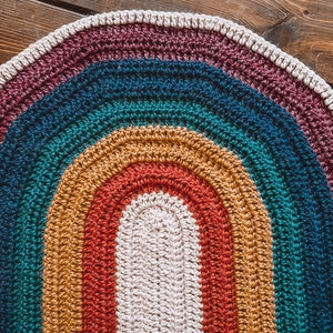 May include: A crocheted oval rug with a rainbow pattern in shades of burgundy, teal, mustard yellow, and red. The center of the rug is a cream color.