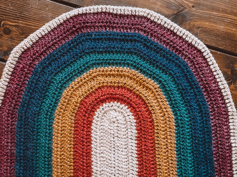 May include: A crocheted oval rug with a rainbow pattern in shades of burgundy, teal, mustard yellow, and red. The center of the rug is a cream color.