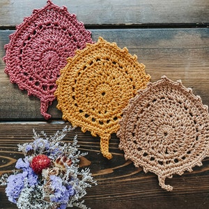 May include: Three crocheted leaf-shaped coasters in shades of pink, orange, and brown. The coasters are arranged in a triangular shape on a wooden surface.