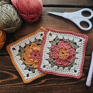 K&ouml;nnte beinhalten: Zwei geh&auml;kelte Quadrate mit floralen Designs in Orange und Rosa, neben Wollkn&auml;ueln in verschiedenen Farben. Eine wei&szlig;e Schere und eine H&auml;kelnadel befinden sich ebenfalls auf einer dunklen Holzoberfl&auml;che, was auf ein Bastelthema hindeutet.