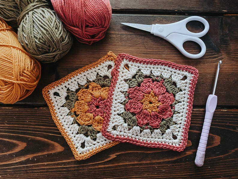 K&ouml;nnte beinhalten: Zwei geh&auml;kelte Quadrate mit floralen Designs in Orange und Rosa, neben Wollkn&auml;ueln in verschiedenen Farben. Eine wei&szlig;e Schere und eine H&auml;kelnadel befinden sich ebenfalls auf einer dunklen Holzoberfl&auml;che, was auf ein Bastelthema hindeutet.