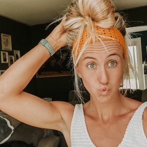 May include: A person with blonde hair in a bun wears an orange crocheted headband. They are wearing a white top and a light green watch. The background features framed pictures and a dark green wall.