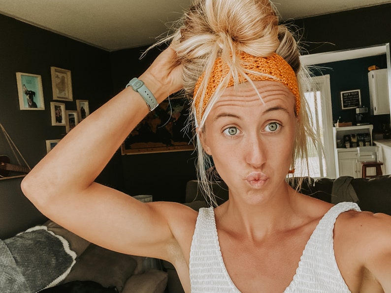 May include: A person with blonde hair in a bun wears an orange crocheted headband. They are wearing a white top and a light green watch. The background features framed pictures and a dark green wall.