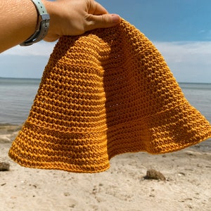 Op de afbeelding: Een gebreide bucket hat in een mosterdgele kleur. De hoed wordt in een hand gehouden tegen een achtergrond van een strand en blauwe lucht.